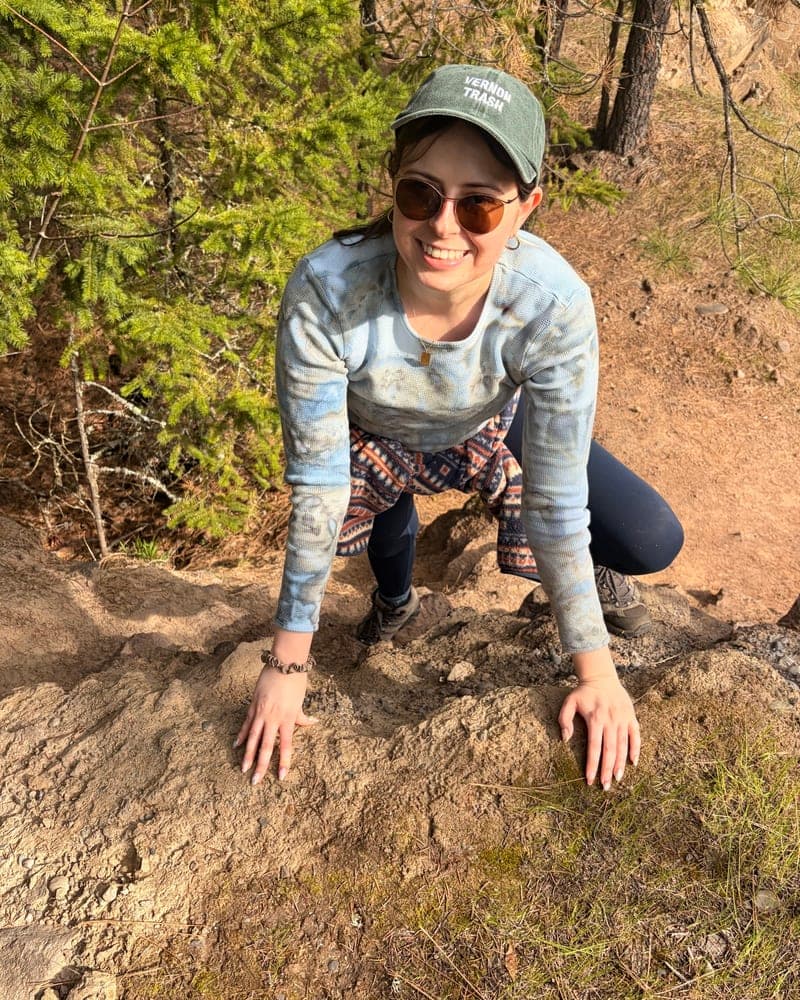 Taylor Villucci outdoors on a sunlit trail, smiling, wearing a Vernon neighborhood cap.